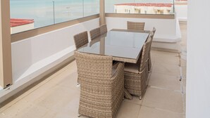 Ett takförsett utomhusområde med bord och stolar, med utsikt över en strand och stadens silhuett.