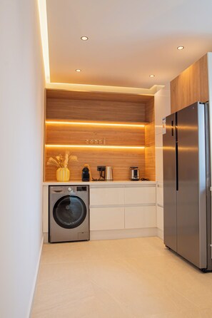 A compact kitchen with a stainless steel refrigerator, a washing machine, and wooden shelves with built-in lighting. - 3-bedroom penthouse, stunning beach front location, minutes to shopping, train, restaurants. (Tangier)