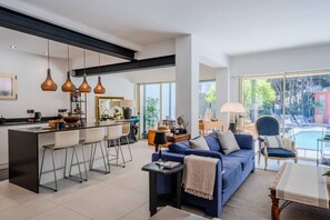 Ein modernes Wohnzimmer mit einem blauen Sofa, einem Couchtisch und einem Esstisch. Es gibt eine Küchenzeile mit einer Bar und Pendelleuchten.