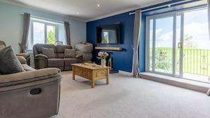 A modern living room with a sofa, a recliner, and a wooden coffee table. There is a TV mounted on the wall and a large window with a balcony view. - The House at Cherry Tree (Kendal)