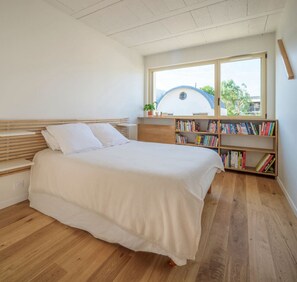 Ein Schlafzimmer mit Bett, Holzboden, einem Bücherregal und einem Fenster mit Blick auf ein kuppelartiges Gebäude.