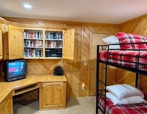 Ein Etagenbett mit rot-weißer Karodecke, ein Holztisch mit Fernseher und ein Bücherregal voller Bücher und DVDs.