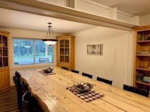 Une salle à manger avec une longue table en bois, des chaises, un lustre et une fenêtre donnant sur les arbres.