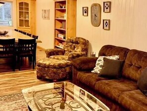 A living room with a brown sofa, patterned armchair, glass coffee table, wooden dining table with chairs, and a bookshelf.