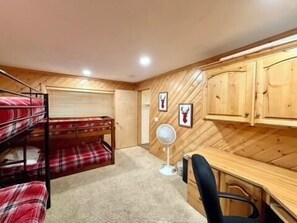 Ein Stockbett mit Karo-Bettwäsche, ein Schreibtisch mit Stuhl, Holzschränke und ein Wandventilator.