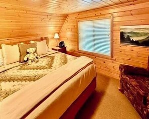 A wooden cabin bedroom with a bed, a nightstand, a lamp, a window with blinds, a painting, and a red chair.
