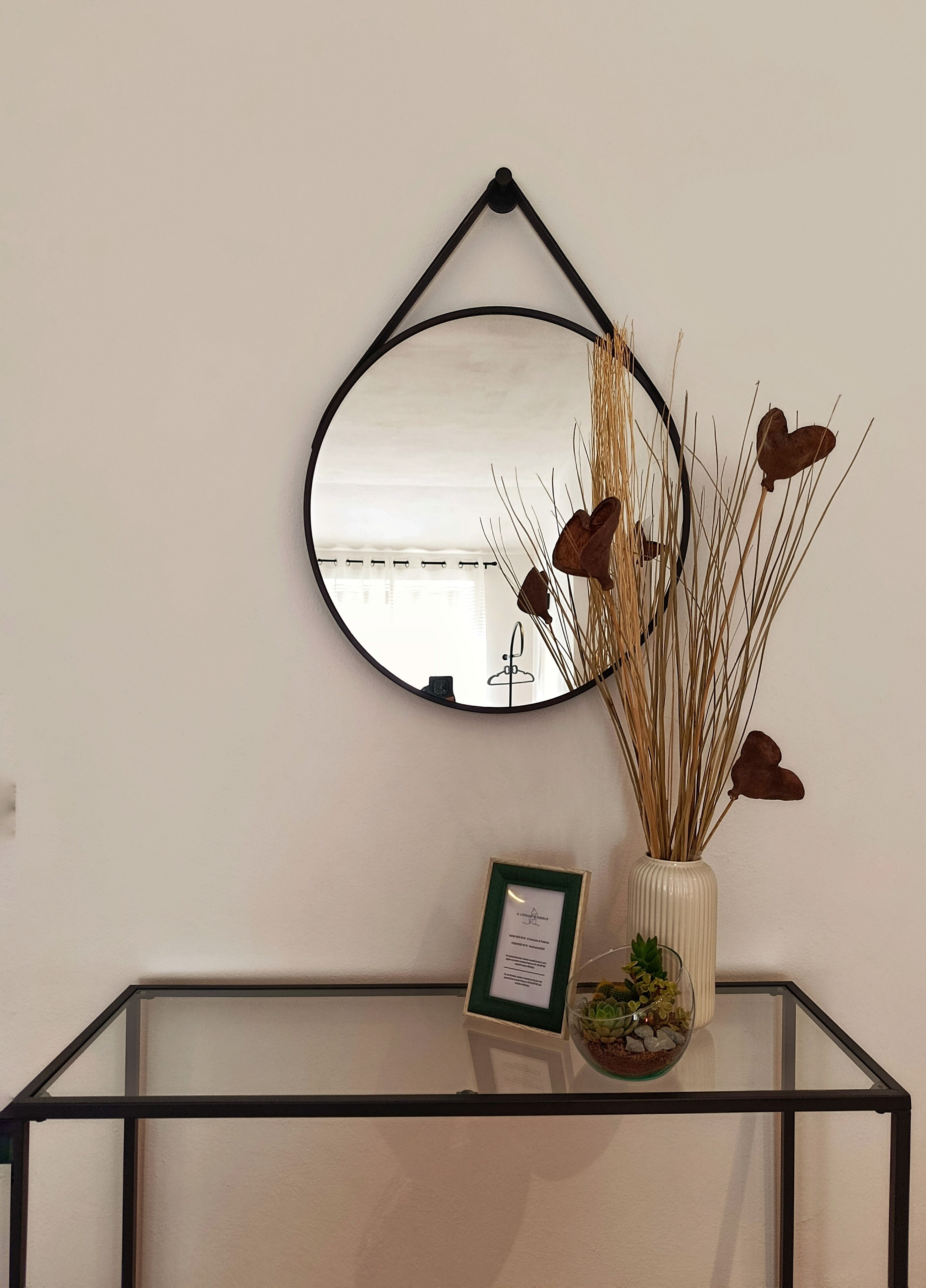 Een spiegel aan de muur, een ingelijst certificaat en een vaas met planten op een glazen tafel.