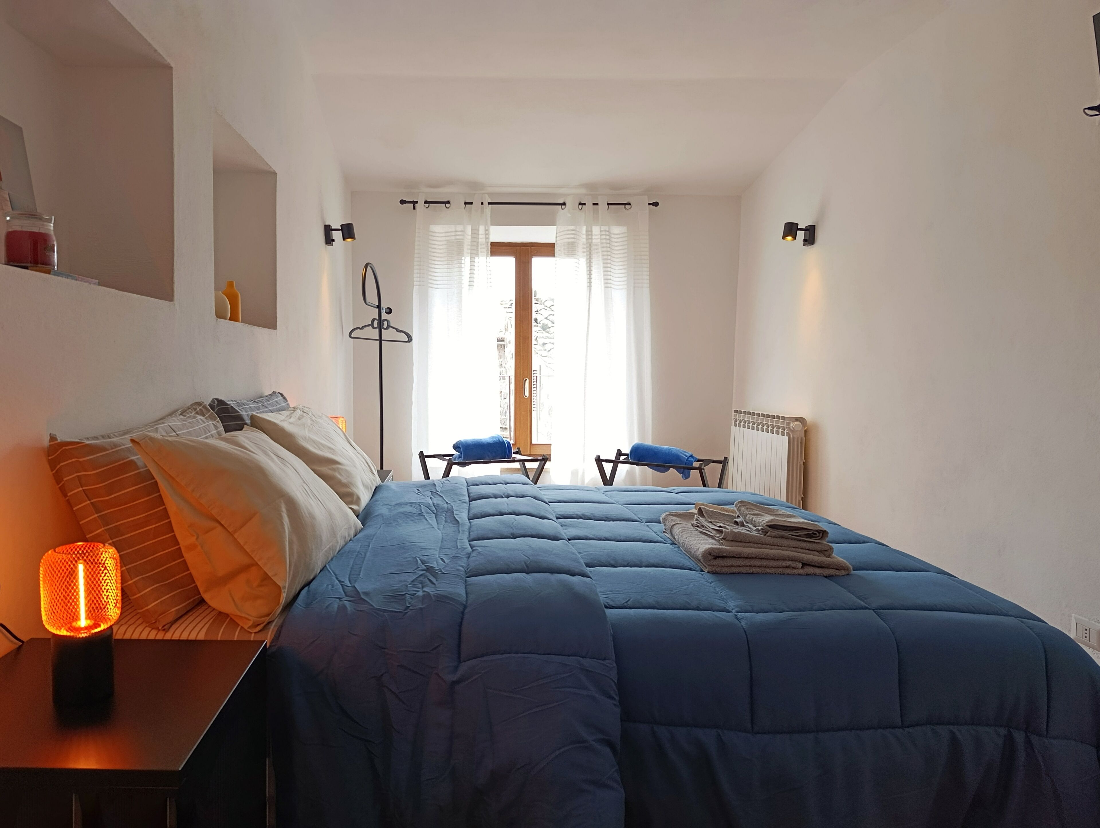 A bedroom with a bed, bedside table, lamp, and a view of the outside through a window.