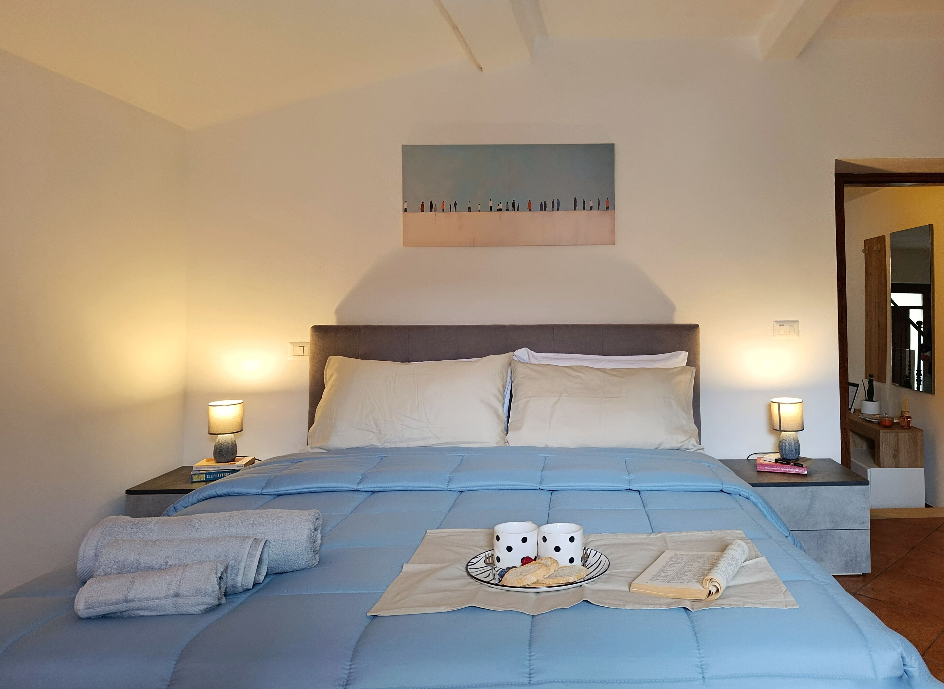 Una cama bien hecha con edredón azul, dos lámparas de noche, una mesita con un libro y un plato de galletas, y un cuadro sobre la cama.