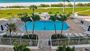 A pool surrounded by palm trees and lounge chairs, with a view of the beach and ocean. - Sky High View at EB& Tennis 1002C-NEW LISTING (Fort Myers Beach)