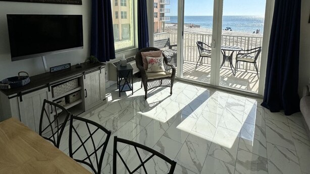 A modern kitchen with a large TV, dining area, and a balcony with a view of the beach. - Sunset Views at EB & Tennis Club 104C (Fort Myers Beach)