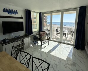 A modern kitchen with a large TV, dining area, and a balcony with a view of the beach. - Sunset Views at EB & Tennis Club 104C (Fort Myers Beach)