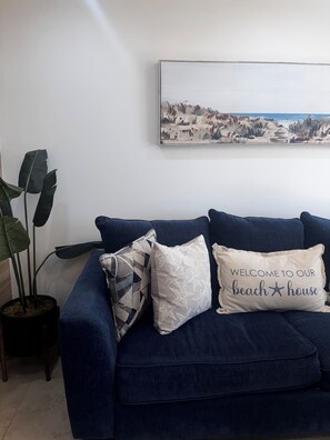 A blue sofa with three decorative pillows, one with a beach house message, and a large framed beach landscape painting on the wall.