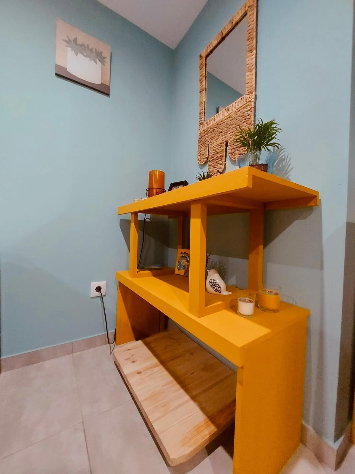 A room with a blue wall, a wooden bench, a yellow console table with a mirror, and a small potted plant.