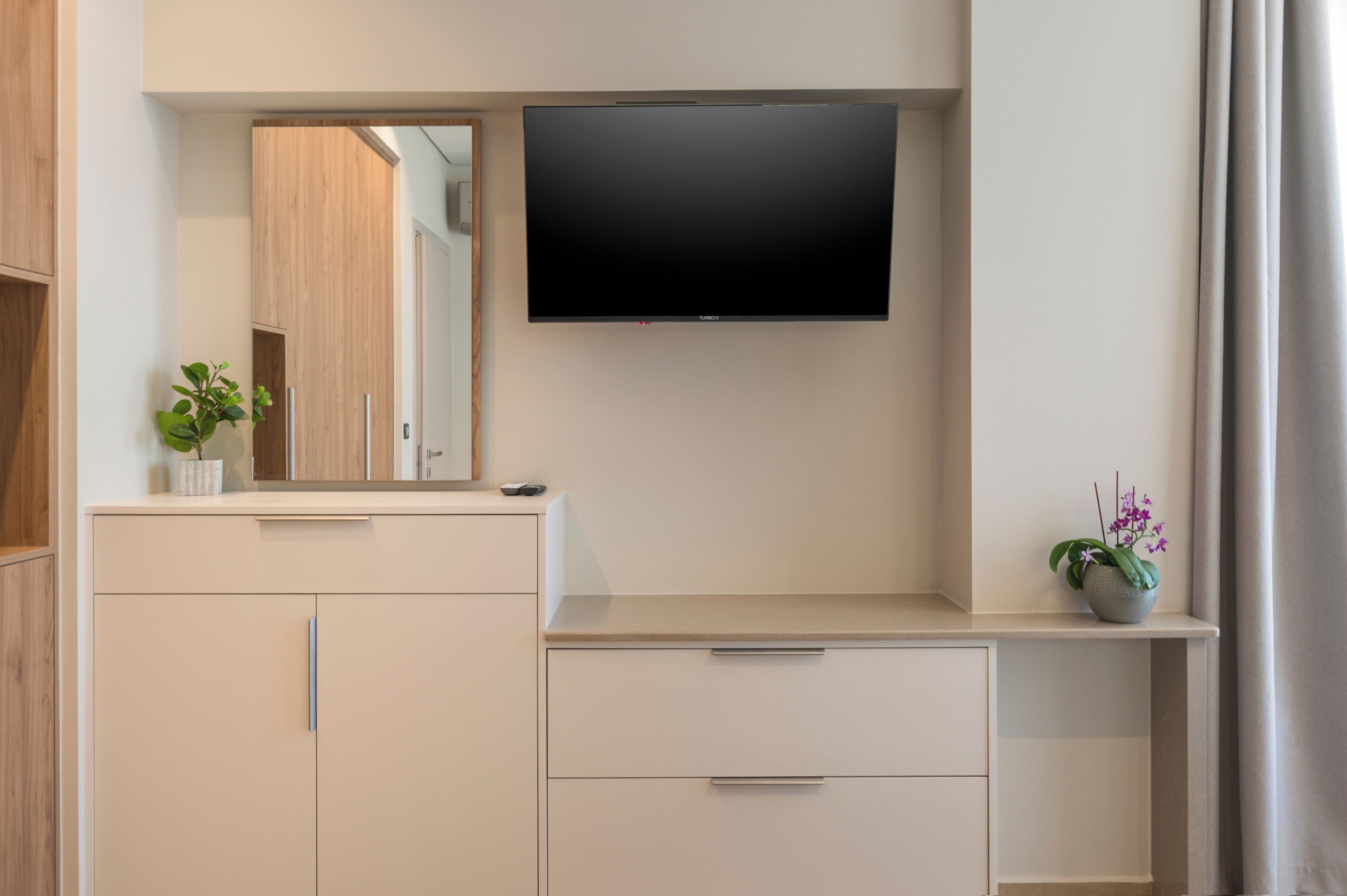 A modern bathroom with a flat-screen TV mounted above a vanity, a mirror, and a potted orchid.