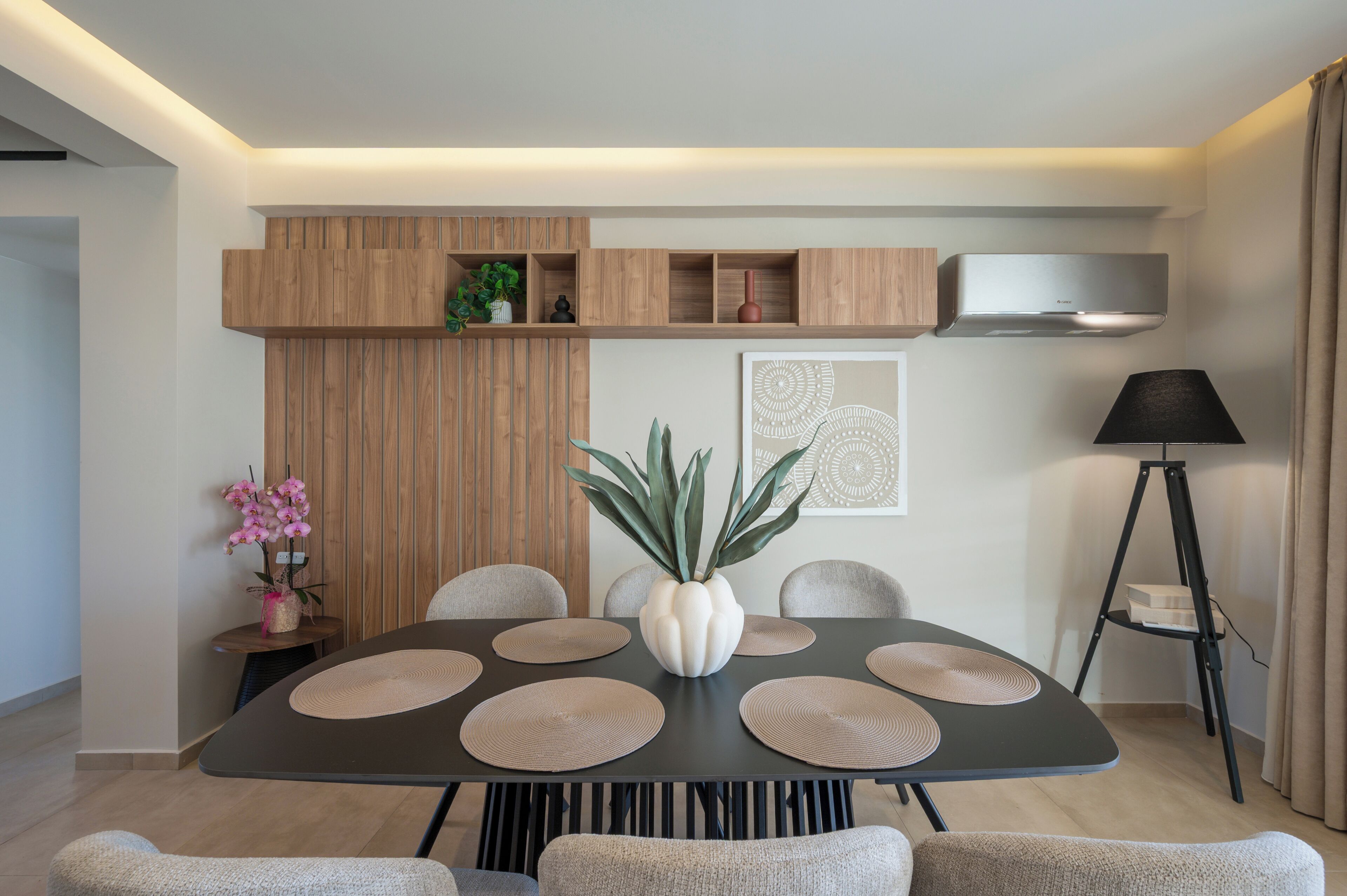 A modern dining area with a round table, multiple chairs, a central vase, and a wall-mounted shelf with decorative items.
