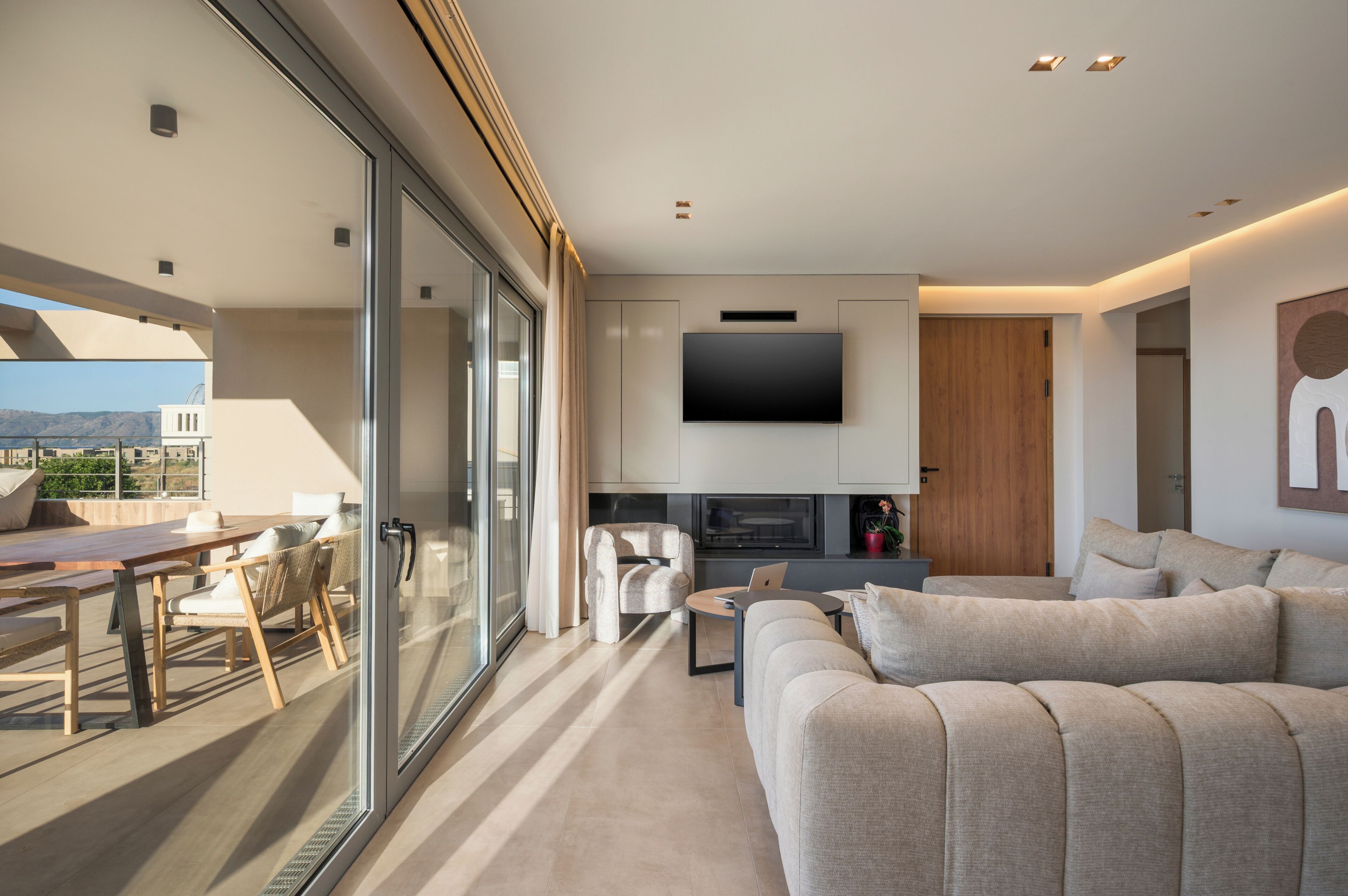 Una sala de estar moderna con un sofá, un televisor montado en la pared, una zona de comedor con mesa y sillas, y un portón corredera de vidrio que da a un espacio exterior.