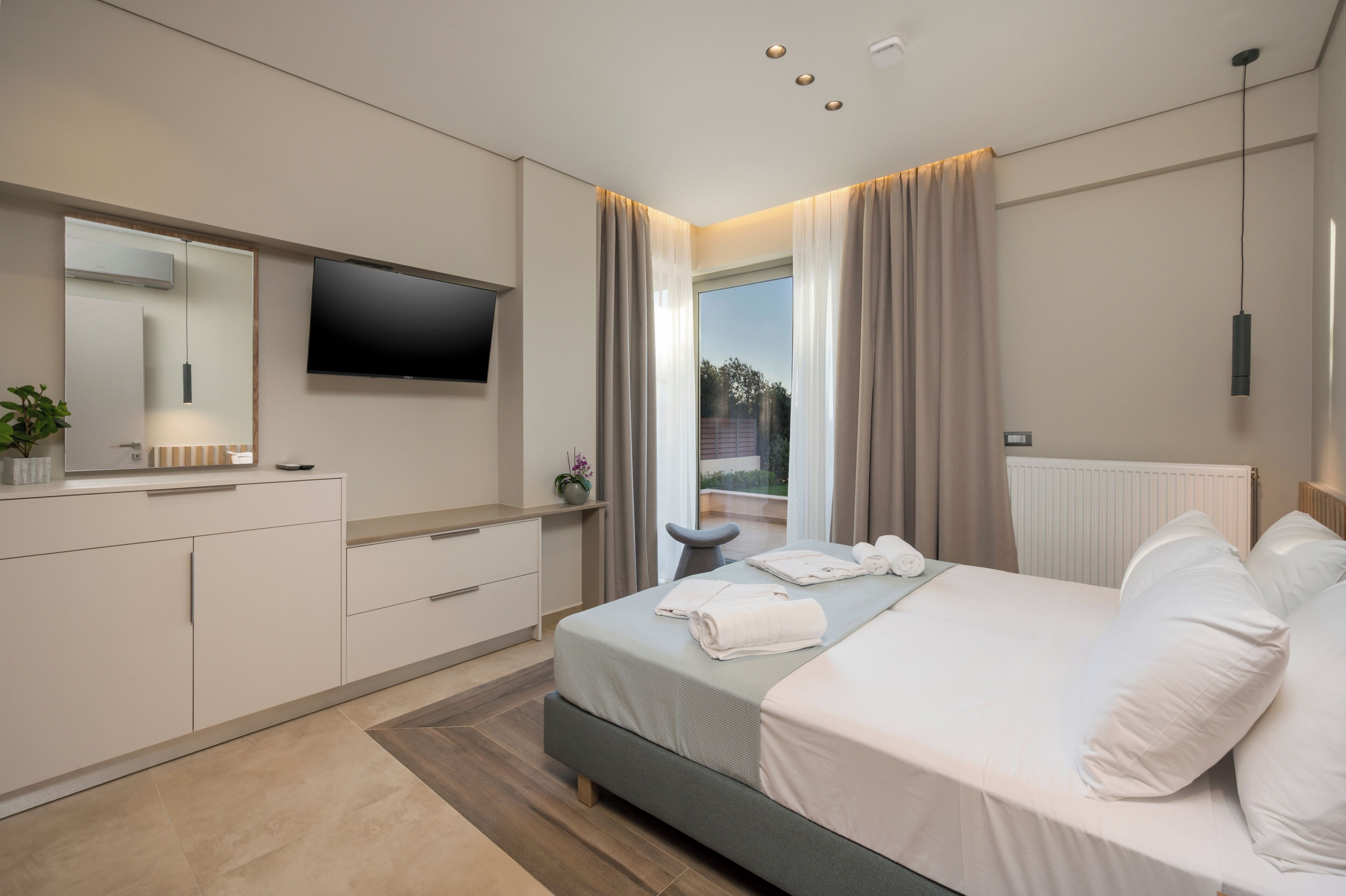 A modern bedroom with a bed, a TV, a desk, and a view of the outdoors.
