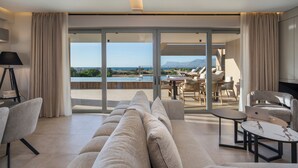A modern living room with a sofa, a coffee table, and a laptop on a table. There is a view of a pool and mountains in the background. - The New Life Houses (Kissamos)