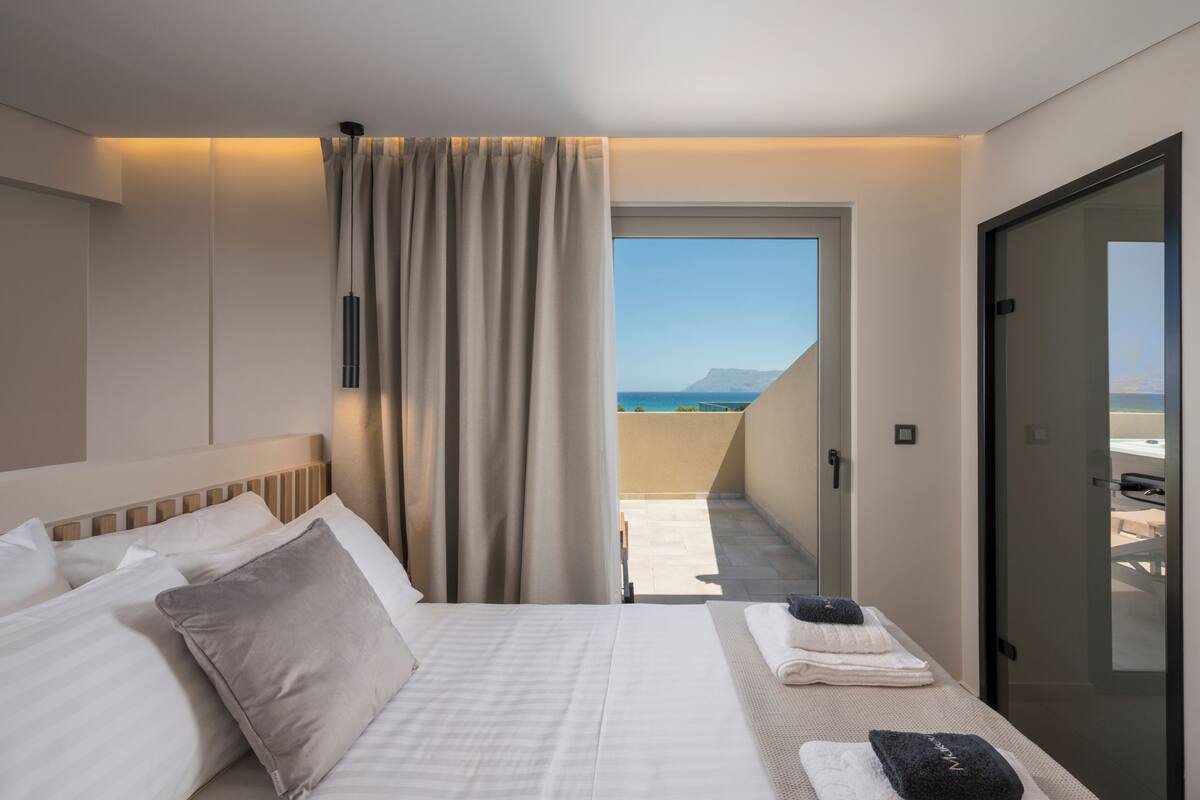 A hotel room with a bed, curtains, a view of the sea, and a door leading to a balcony.