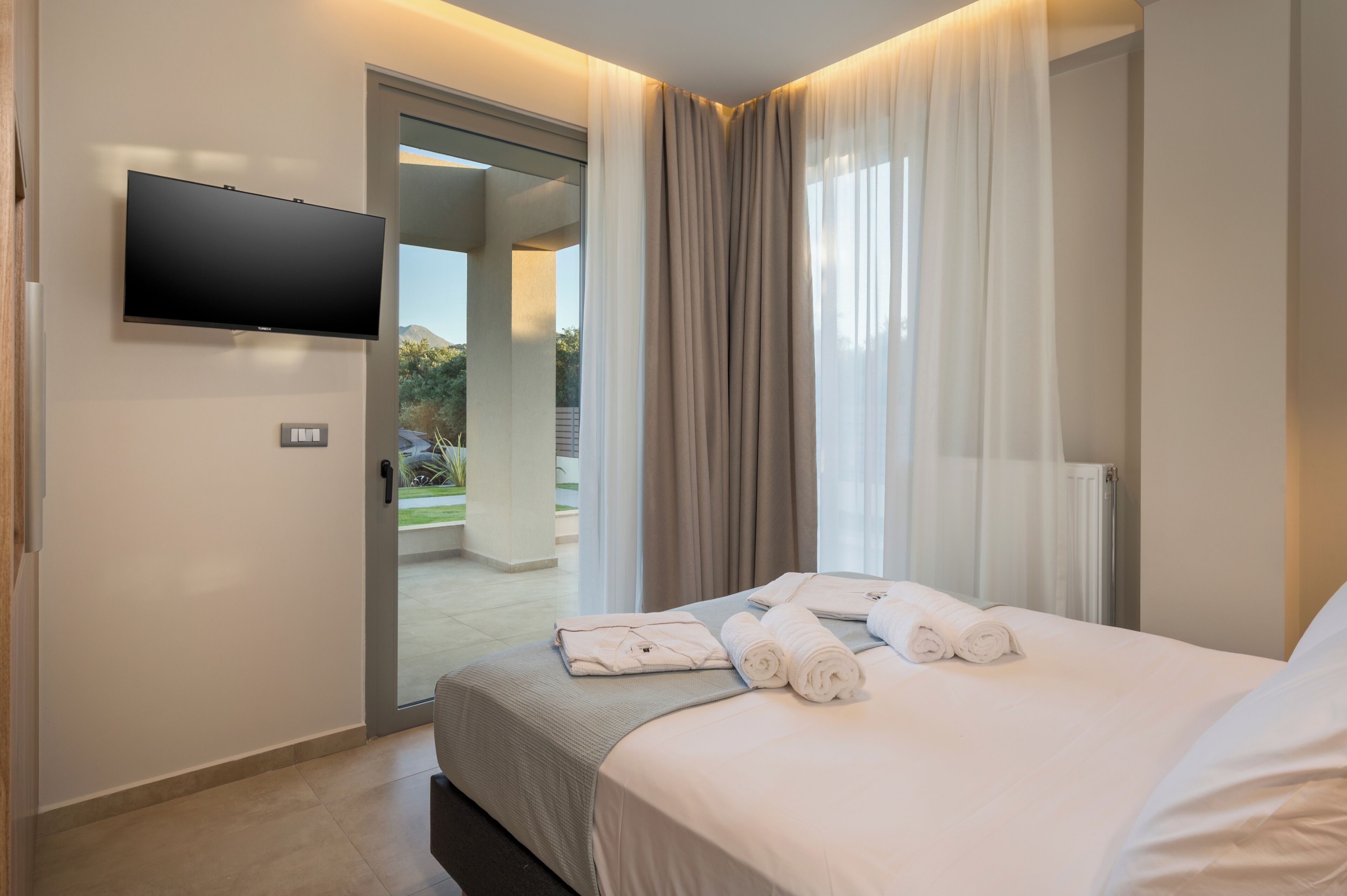 A hotel room with a bed, a flat-screen TV, a sliding glass door leading to a balcony, and a thermostat on the wall.