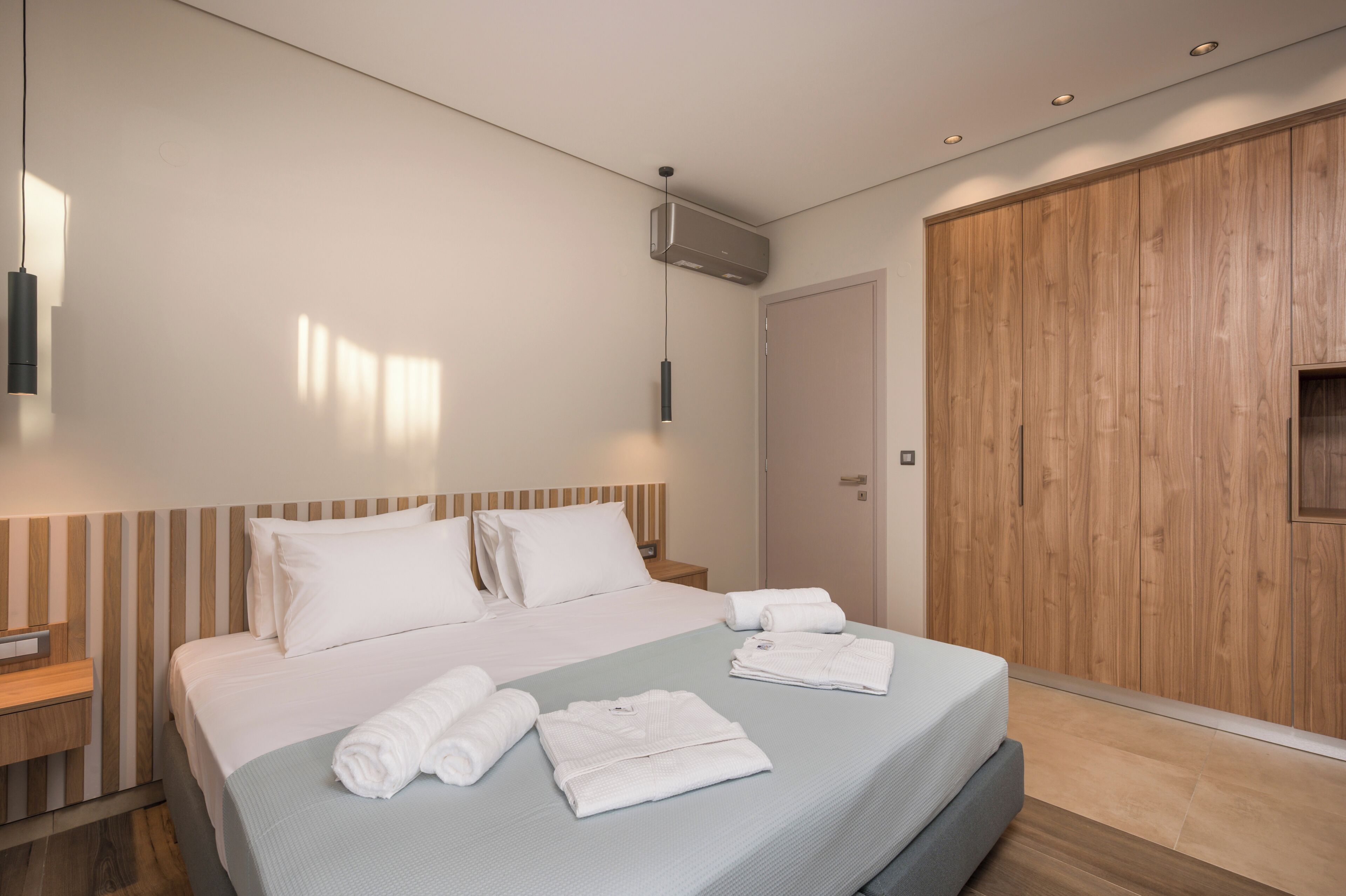 Moderni hotellihuone, jossa on suuri sänky, puinen vaatekaappi, yöpöytä ja seinälle asennettu ilmastointilaite.