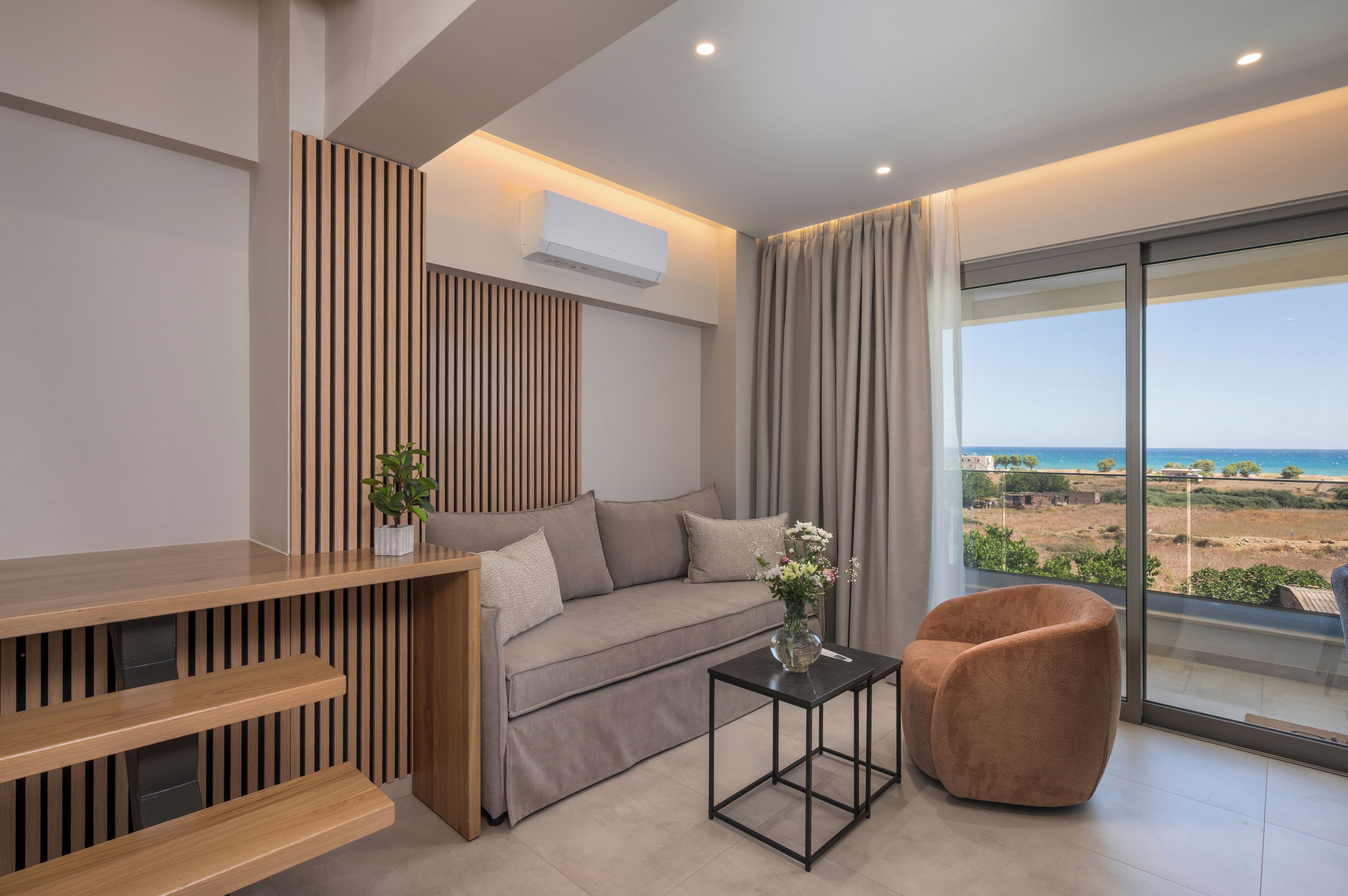 Ein modernes Wohnzimmer mit einer Couch, einem kleinen Tisch mit einer Vase, einem Sessel und Blick aufs Meer.