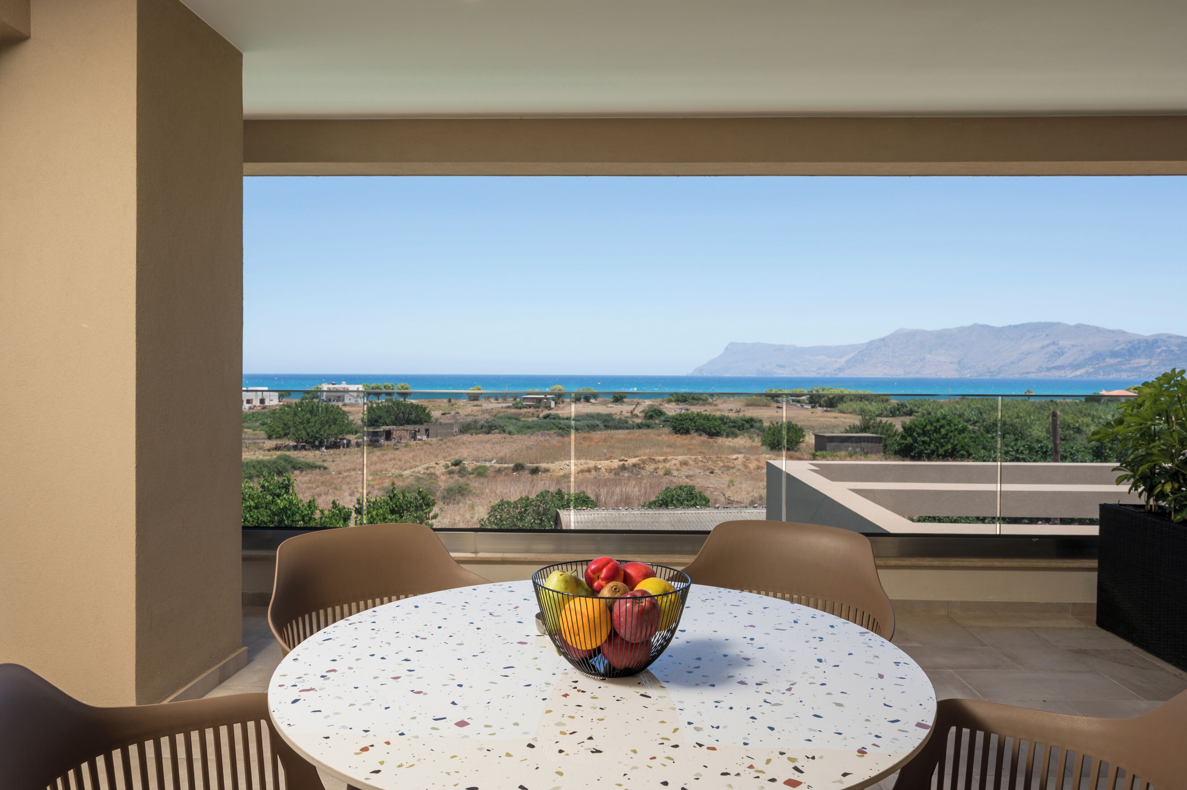 Un balcón con mesa y sillas, un frutero con fruta y vistas al mar y a las montañas.