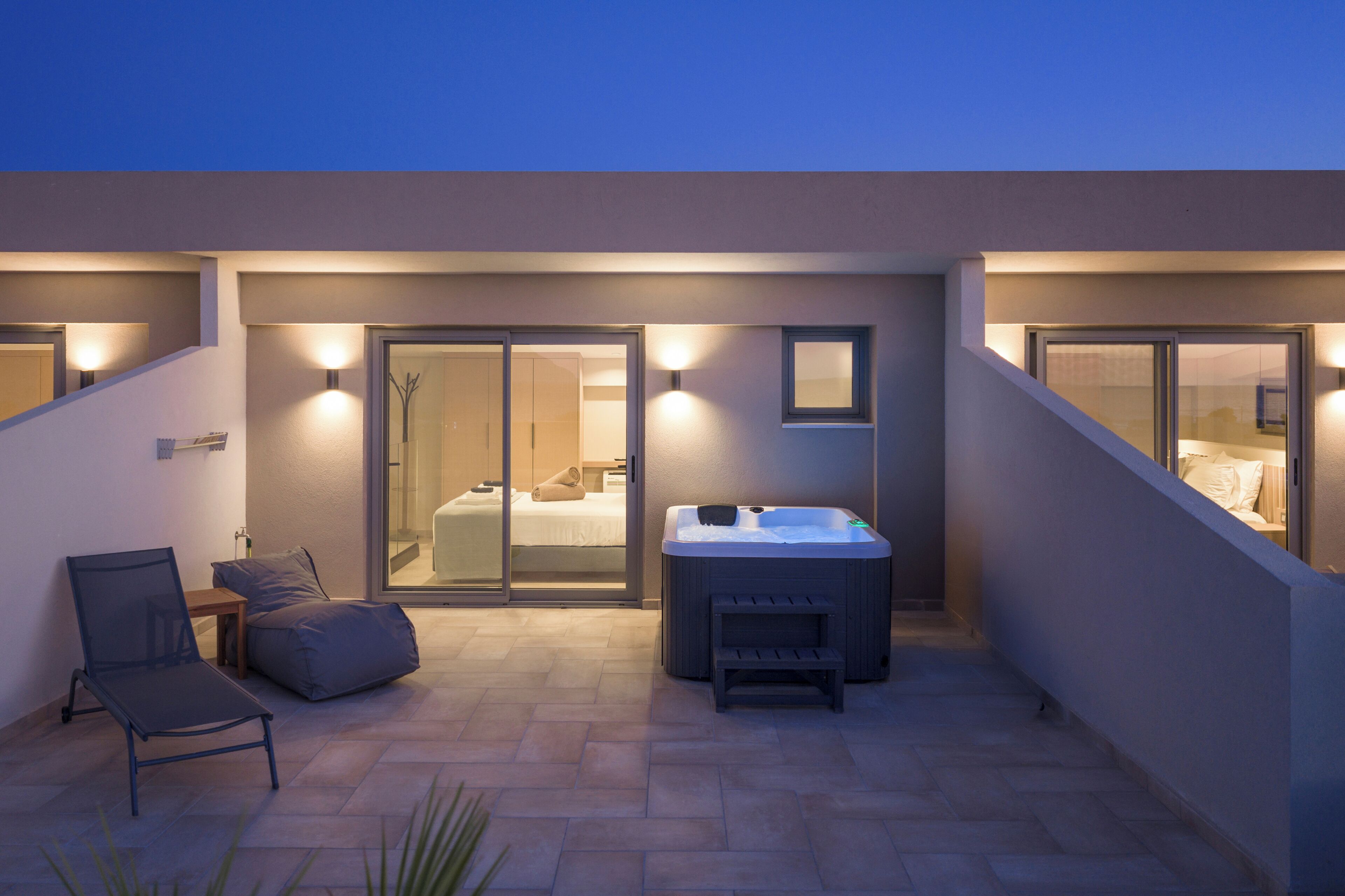 A modern outdoor patio with a hot tub, seating area, and sliding glass doors leading to a bedroom.