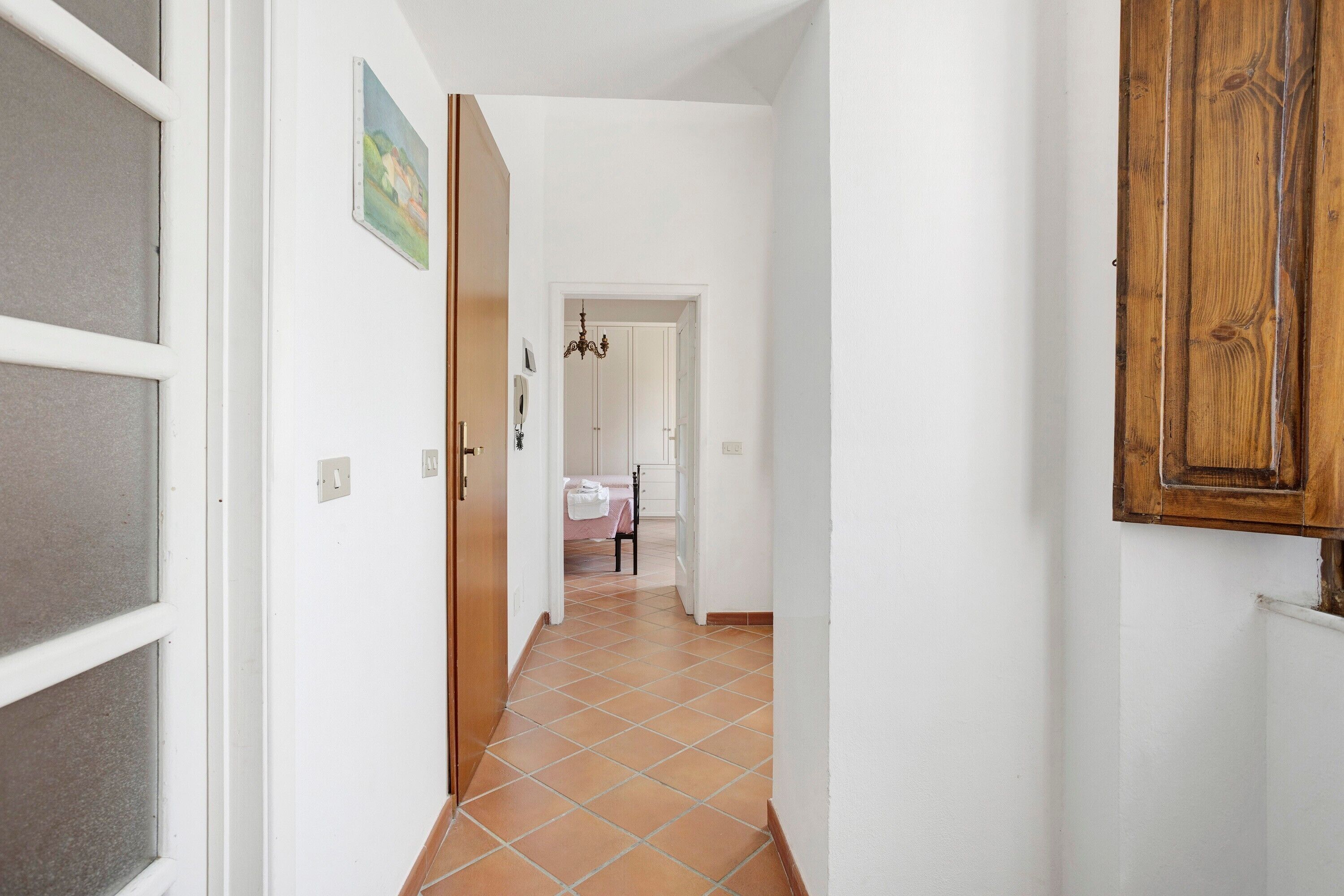 A hallway with a tiled floor, white walls, a wooden door, and a small painting on the wall.