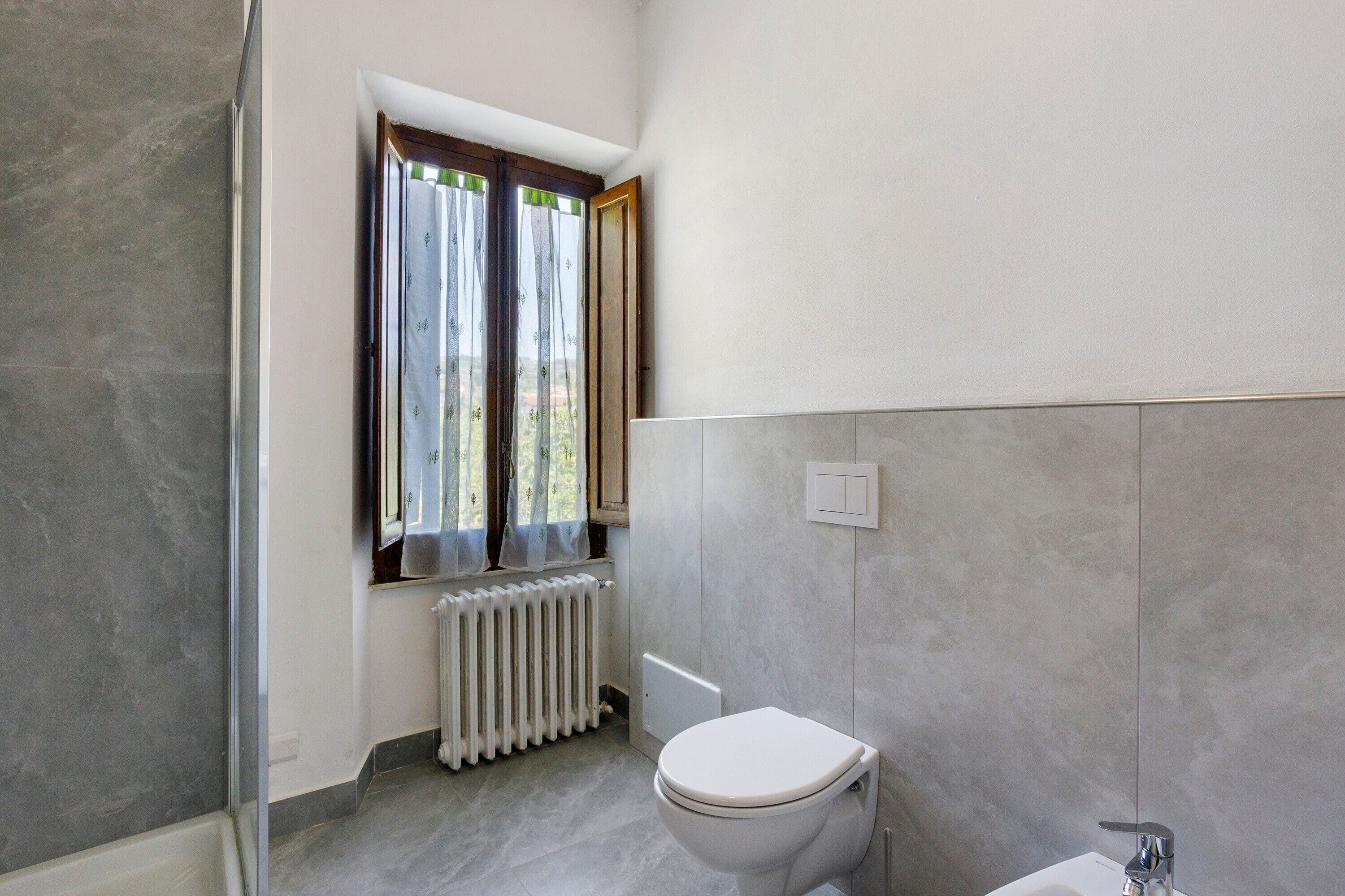 Een badkamer met een toilet, een raam met gordijnen, een radiator en een douche.