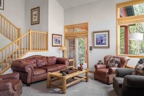 A living room with a brown leather sofa, a wooden coffee table, and a fireplace. There is a staircase with wooden railings and a door leading outside. - Summer Retreat by InvitedHome | BBQ, Hot Tub, Dog Friendly, 5 Min to Downtown (Breckenridge)