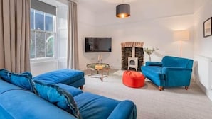 Un soggiorno moderno con un divano blu, un pouf rosso, un tavolino con una lampada e una televisione a parete.