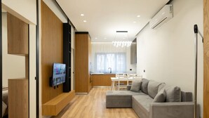 Una sala de estar moderna con un sofá seccional gris, piso de madera, un televisor de pantalla plana montado en la pared y una cocina al fondo.