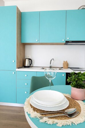 Une cuisine avec des meubles de couleur turquoise, un plan de travail blanc, une machine à café, un évier et un coin repas avec une table mise pour deux personnes.