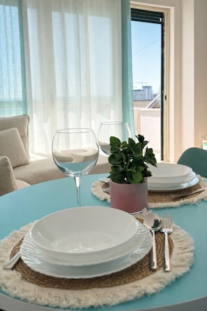 Une table à manger dressée avec des assiettes, des couverts et un verre d’eau, offrant une vue sur la ville par la fenêtre.