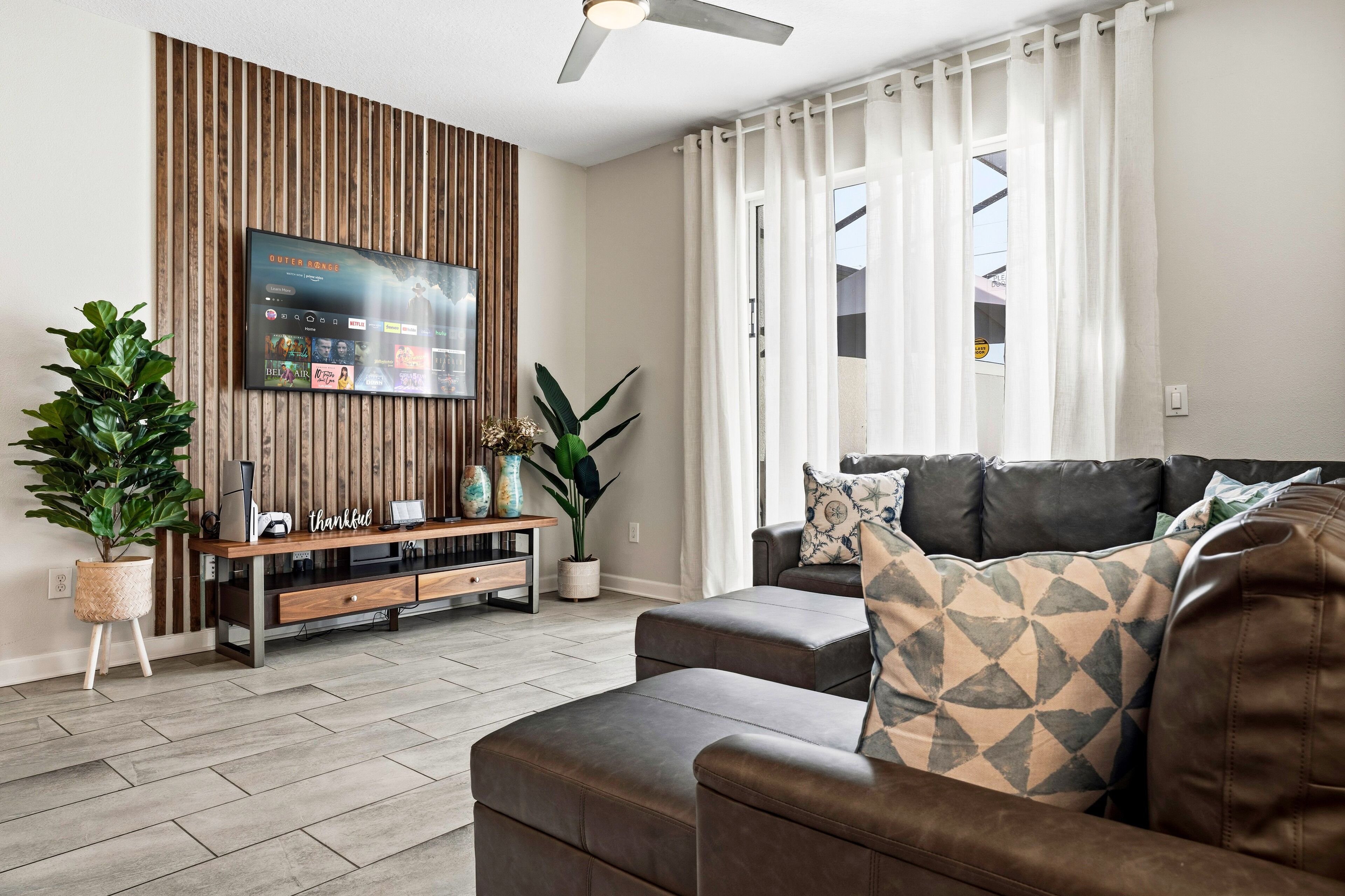 A modern living room with a flat-screen TV mounted on a wooden panel wall, a leather sofa, a coffee table, and a potted plant.