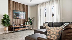 A modern living room with a flat-screen TV mounted on a wooden panel wall, a leather sofa, a coffee table, and a potted plant. - Spacious 4-Bedroom Townhome with PS5, Private Pool & BBQ Grill at Storey Lake (Kissimmee)