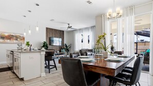 A modern kitchen with a central island, dining area with a wooden table and chairs, and a living area with a sofa and TV. - Spacious 4-Bedroom Townhome with PS5, Private Pool & BBQ Grill at Storey Lake (Kissimmee)