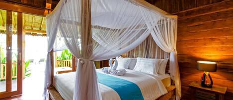 A four-poster canopy bed with white linens and a teal throw blanket, set in a wooden room with a wooden floor and a nightstand with a lamp.