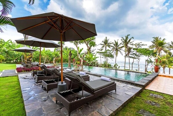 A poolside area with lounge chairs and umbrellas, surrounded by lush greenery and a view of the ocean. - Bali Natha Beach Front (Cemagi)