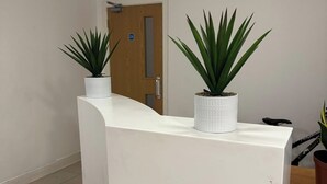 A reception desk with two potted plants, a wooden door, and a tiled floor. - Heathrow Airport - Apt - Stay 3 - Great Location (Hounslow)