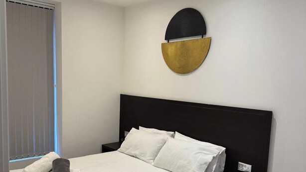 A neatly made bed with white linens, a dark headboard, and a small bedside table with a lamp. - Heathrow Airport - Apt - Stay 3 - Great Location (Hounslow)