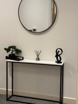 A white console table with a round mirror above it, a bonsai tree, a small vase with reed sticks, and a black abstract sculpture. - Heathrow Airport - Apt - Stay 3 - Great Location (Hounslow)