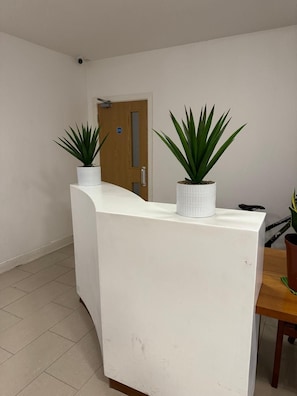 A reception desk with two potted plants, a wooden door, and a tiled floor. - Heathrow Airport - Apt - Stay 3 - Great Location (Hounslow)