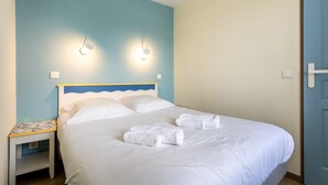 A neatly made bed with white linens and towels, a nightstand with a small table, and two wall-mounted light fixtures. - La Parenthèse Bleue – Sea View and Pool (Fouesnant)