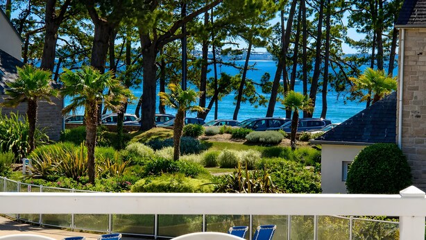 A terrace with a view of the sea, surrounded by trees and parked cars. - La Parenthèse Bleue – Sea View and Pool (Fouesnant)
