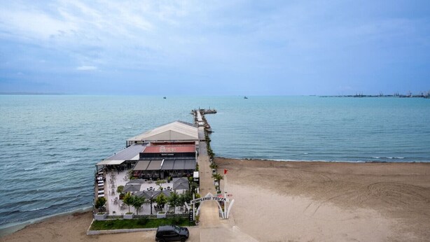 A coastal hotel with a long pier extending into the sea, a sandy beach, and a clear sky.