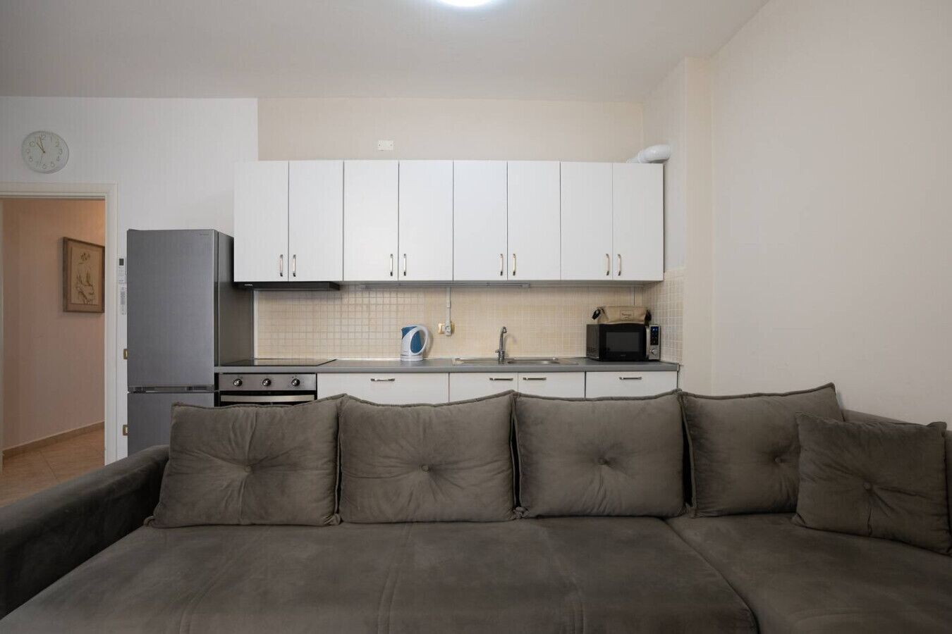A compact living space with a kitchenette, a sofa with cushions, and a clock on the wall.