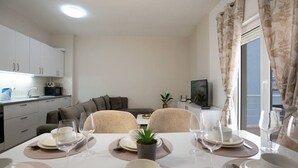 A dining area with a table set for four, a sofa, a television, and a balcony with curtains.